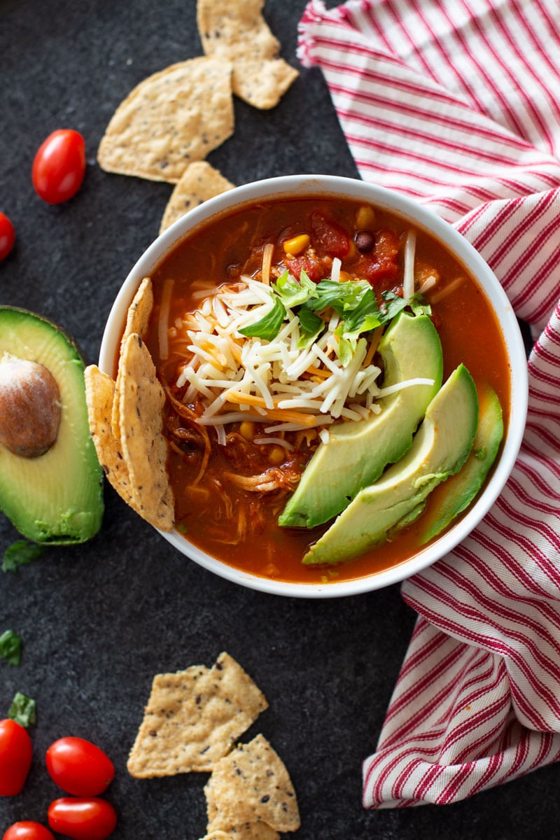 White bowl containing tortilla soup topped with shredded cheese, avocado and cilantro.
