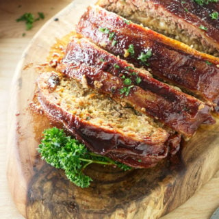 smoked meatloaf wrapped in bacon and brushed with bbq sauce sitting on wood cutting board with red and white towel and parsley