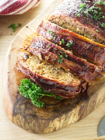 smoked meatloaf wrapped in bacon and brushed with bbq sauce sitting on wood cutting board with red and white towel and parsley