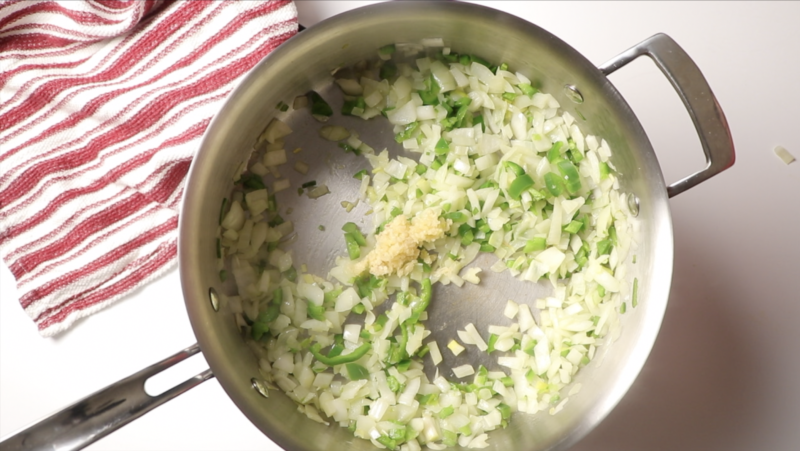 chopped onions jalapenos and garlic in a large saute pan with a red and white towel on white table