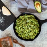 Skillet containing avocado pasta, wine, Parmesan cheese, and pepper on table.