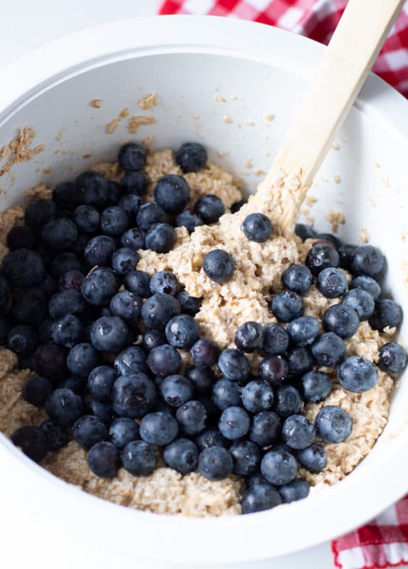 White bowl containing oatmeal mixture topped with fresh blueberries, wooden spoon in bowl.
