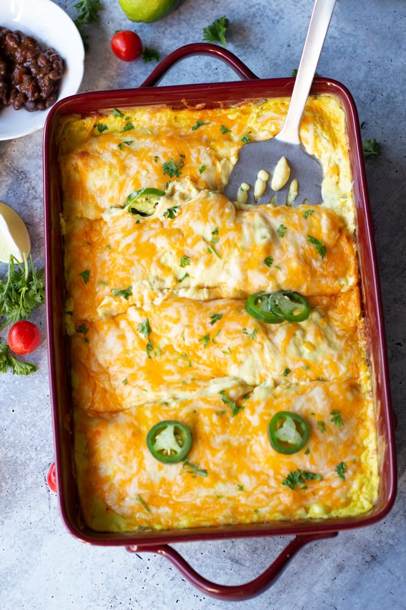 Spatula in a casserole dish removing chicken enchiladas topped with a creamy cheese sauce.