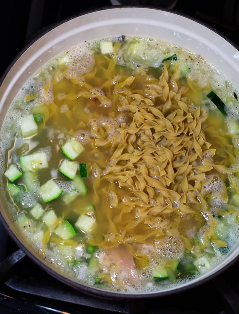Raw egg noodles being added to a stockpot of homemade chicken vegetable soup.