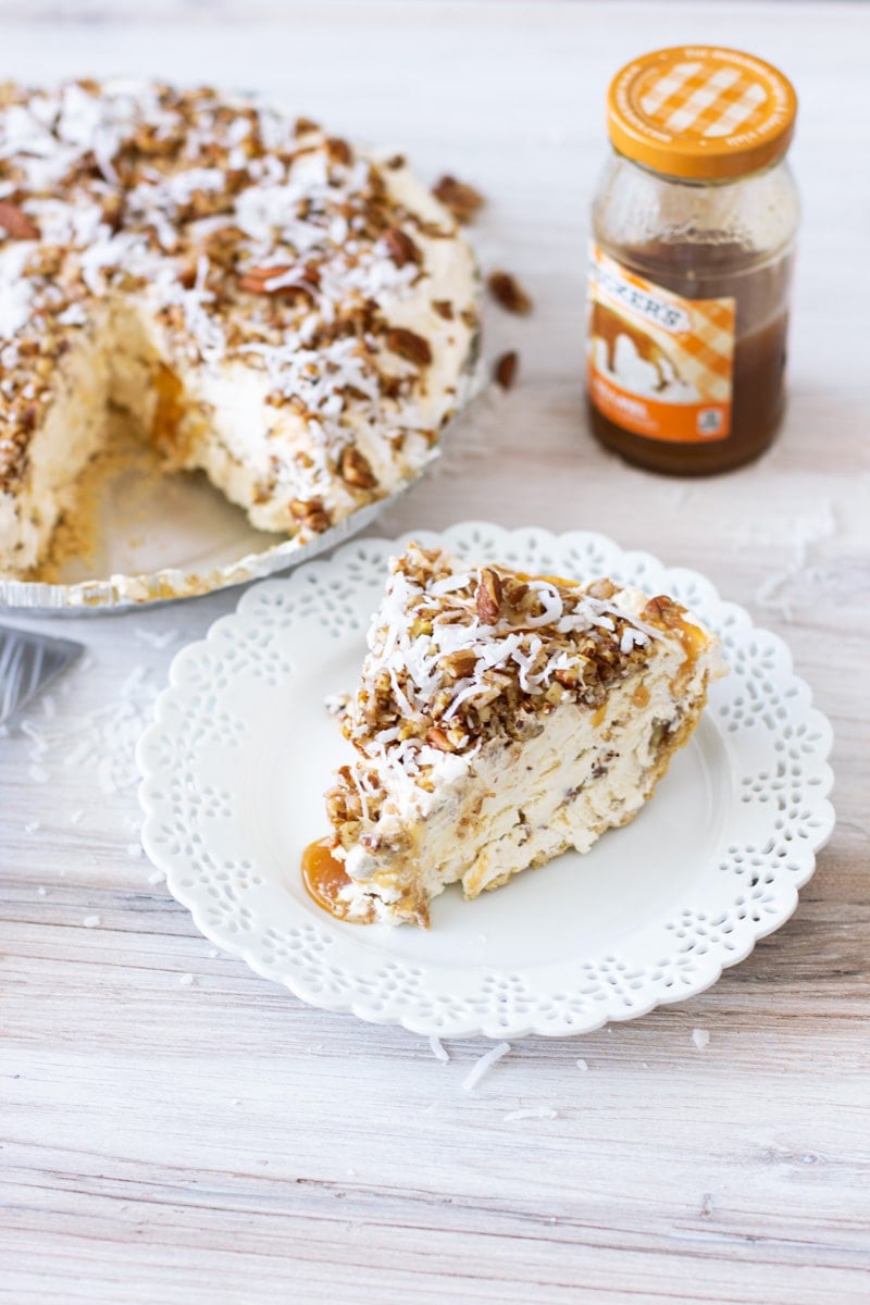 Plate containing a piece of pecan pie topped with pecans and coconut flakes, pie on table in background.