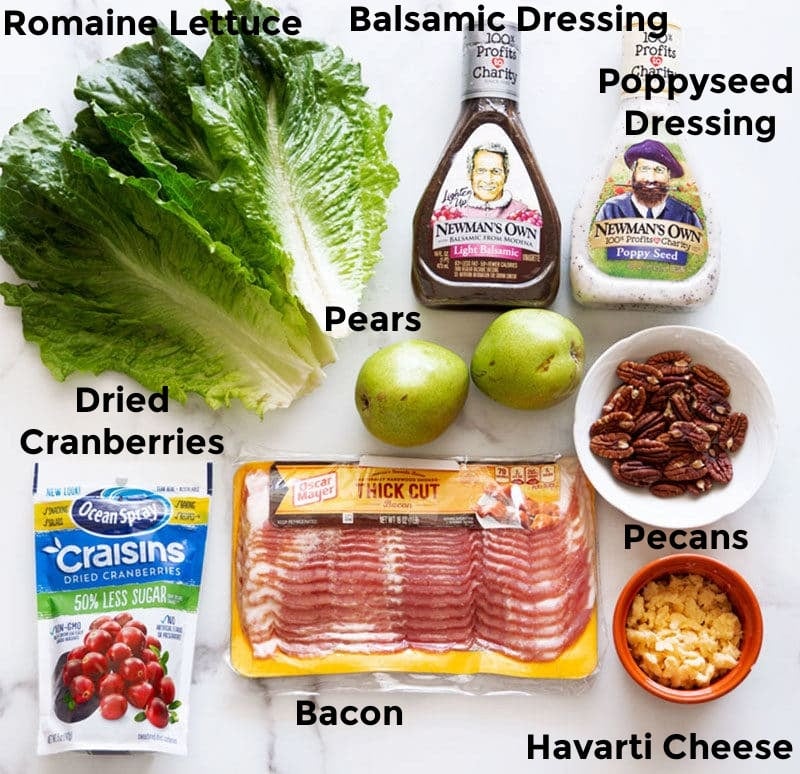 Lettuce, dresiing, pears, bacon, cheese, nuts, and cranberries on a counter. 