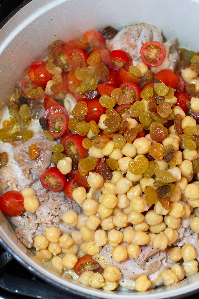 Dutch oven containing chickpeas, raisins, tomatoes covering chicken pieces. 