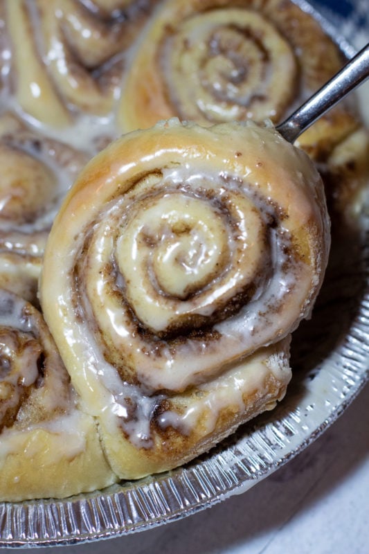 Close up of cinnamon rolls covered in vanilla icing, other cinnamon rolls on platter. 