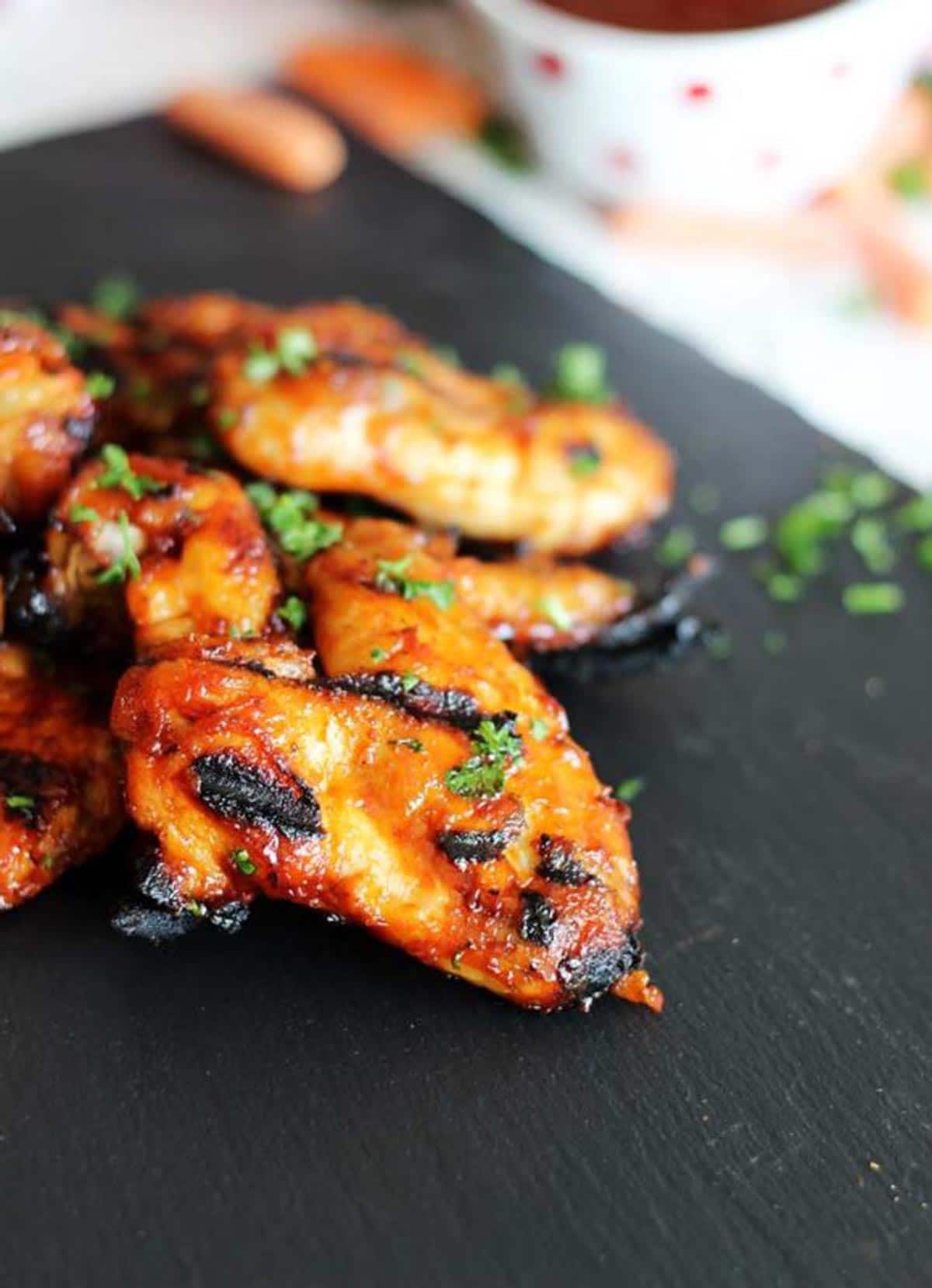 Slate plate featuring 4 grilled chicken wings topped with parsley, BBQ sauce and carrots in the background.