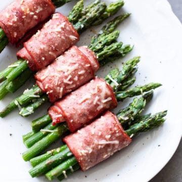 White plate containing asparagus wrapped in bacon topped with Parmesan.