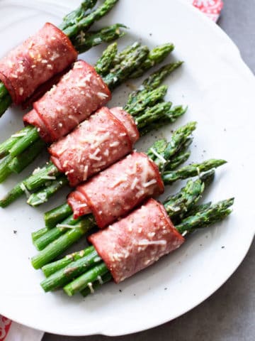 White plate containing asparagus wrapped in bacon topped with Parmesan.