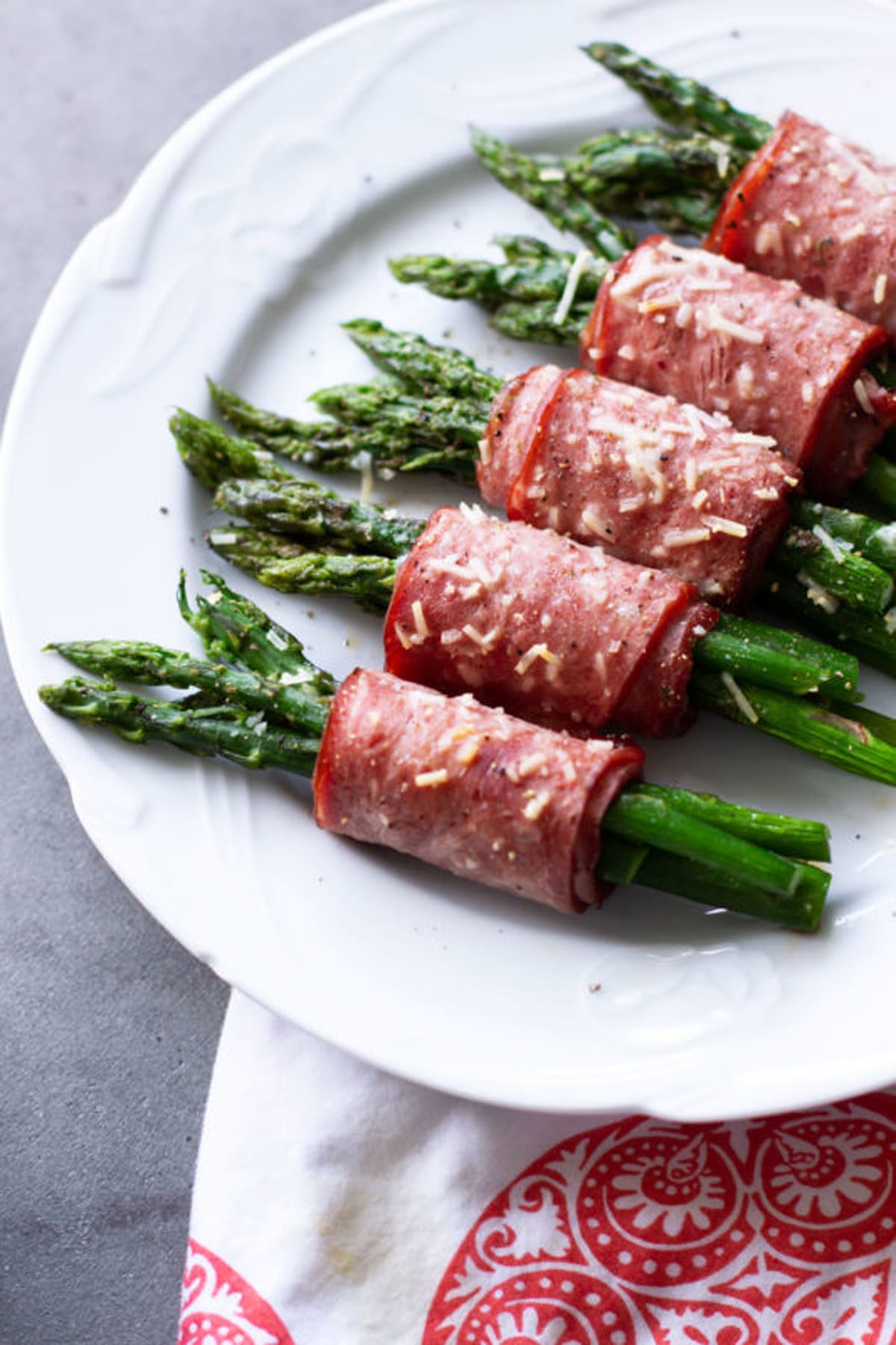 White plate containing asparagus wrapped in bacon topped with Parmesan. 