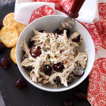 White bowl of chicken salad on a black serving tray, bread, crackers and grapes on serving dish.