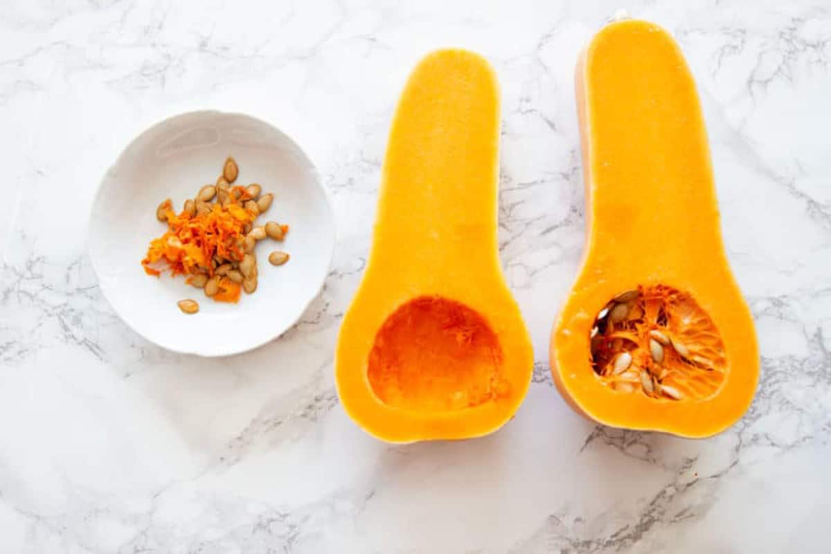 Butternut squash cut in half lengthwise sitting on a table, pumpkin seeds in a bowl.