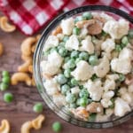 Bowl containing frozen peas, cashew halves, cauliflower, onion and mayonnaise topped with salt and pepper.