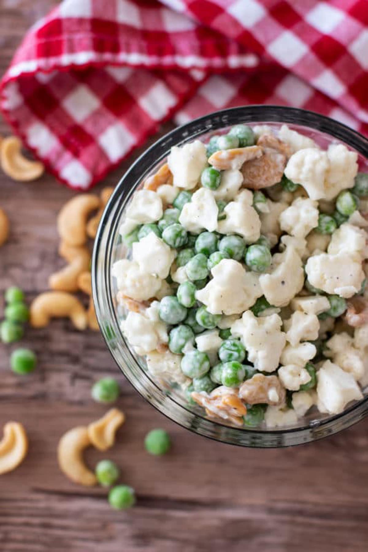 Peas, cauliflower and cashews salad in a glass bowl sitting on a red and white gingham napkin, scattered peas and cashew nuts on table.