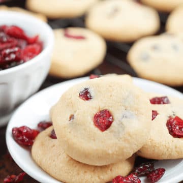 White bowl containing 3 Cranberry Cookies, cranberries and cookies on table.
