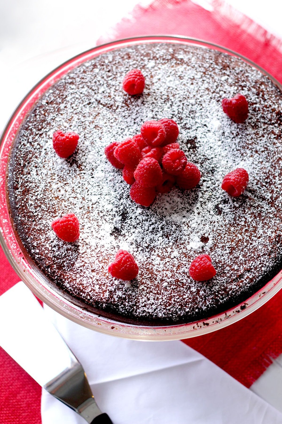 Flourless Chocolate Torte topped with powdered sugar and raspberries sitting on a cake dish, pie server on side.