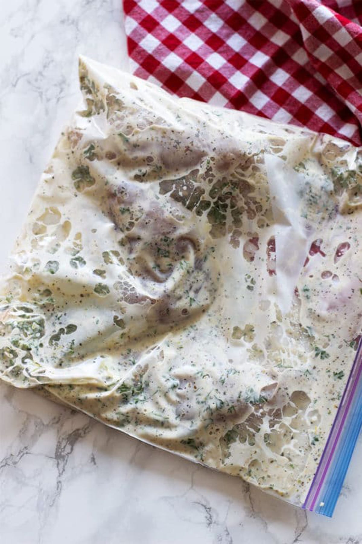 Greek marinated chicken marinating in a Ziplock baggie sitting on a white marble table, red and white gingham napkin on table.