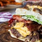 A Juicy Lucy bacon burger cross section, sitting on a wood cutting board, cheese spilling out the middle with bacon, onion, tomato, lettuce, with sweet potato fries in background