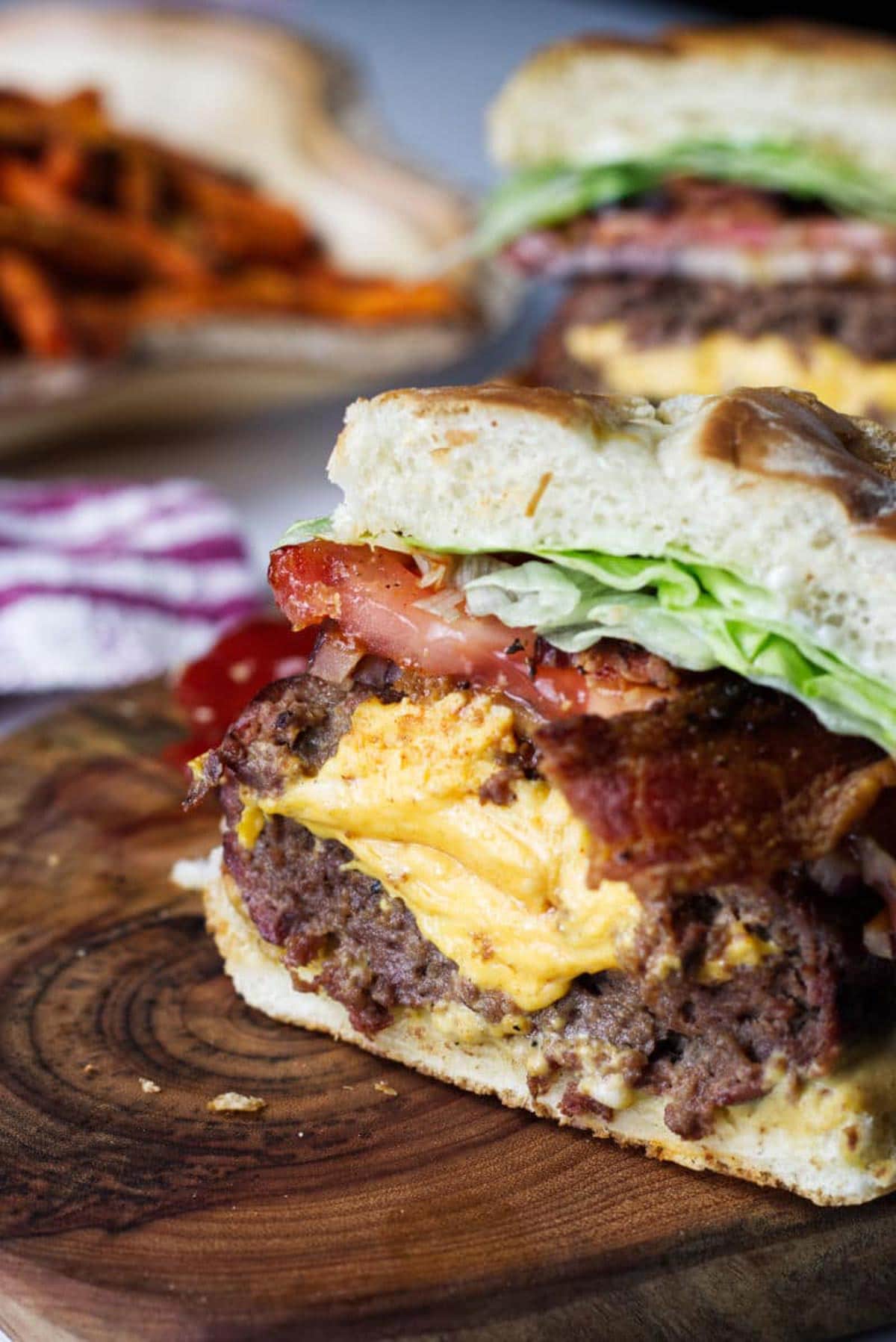 A Juicy Lucy burger cross section, sitting on a wood cutting board, cheese spilling out the middle with bacon, onion, tomato, lettuce, with sweet potato fries in background