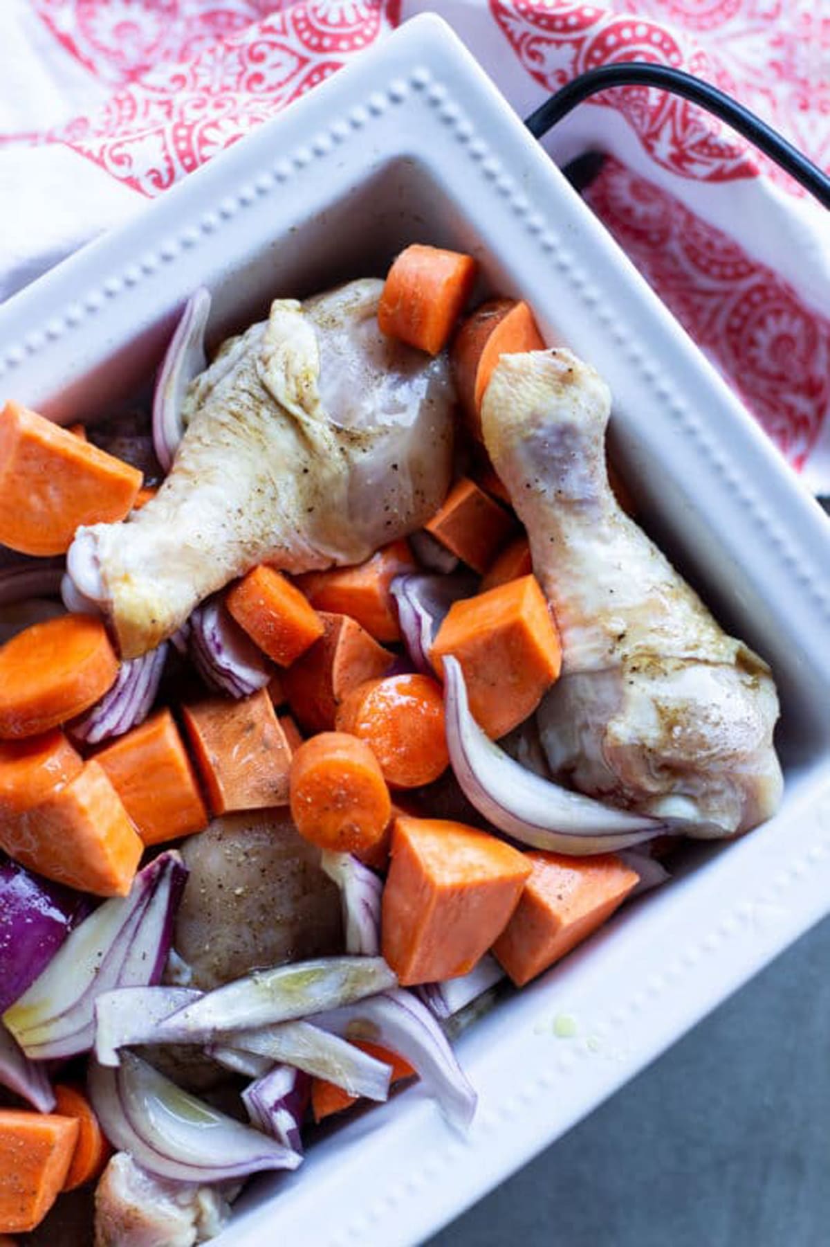 White dish containing seasoned drumsticks with sweet potatoes, carrots, and red onions drizzled with olive oil.
