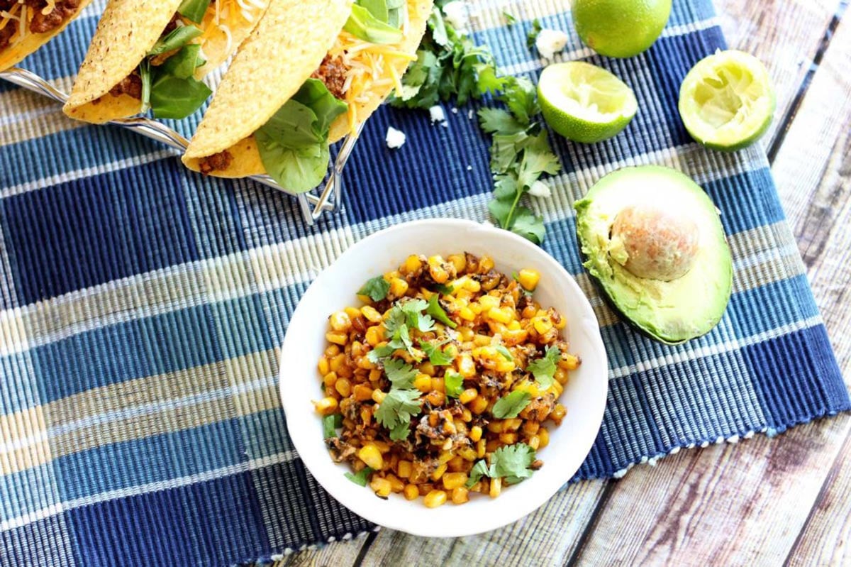 White bowl containing Mexican Street Corn sitting on a blue tablecloth, tacos, cheese, avocado and limes on table.