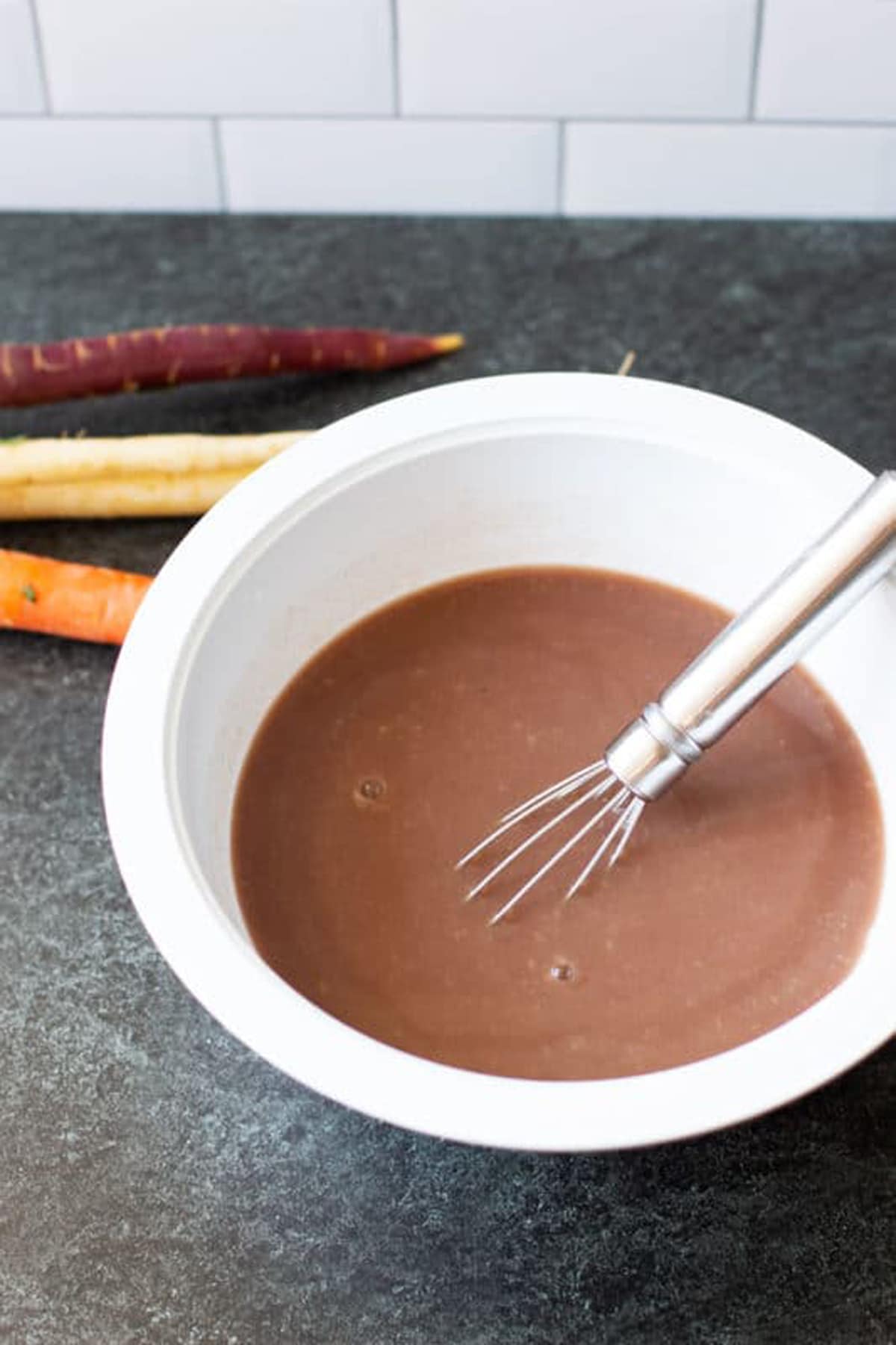 White bowl containing gravy, spatula in bowl. Carrots on counter. 