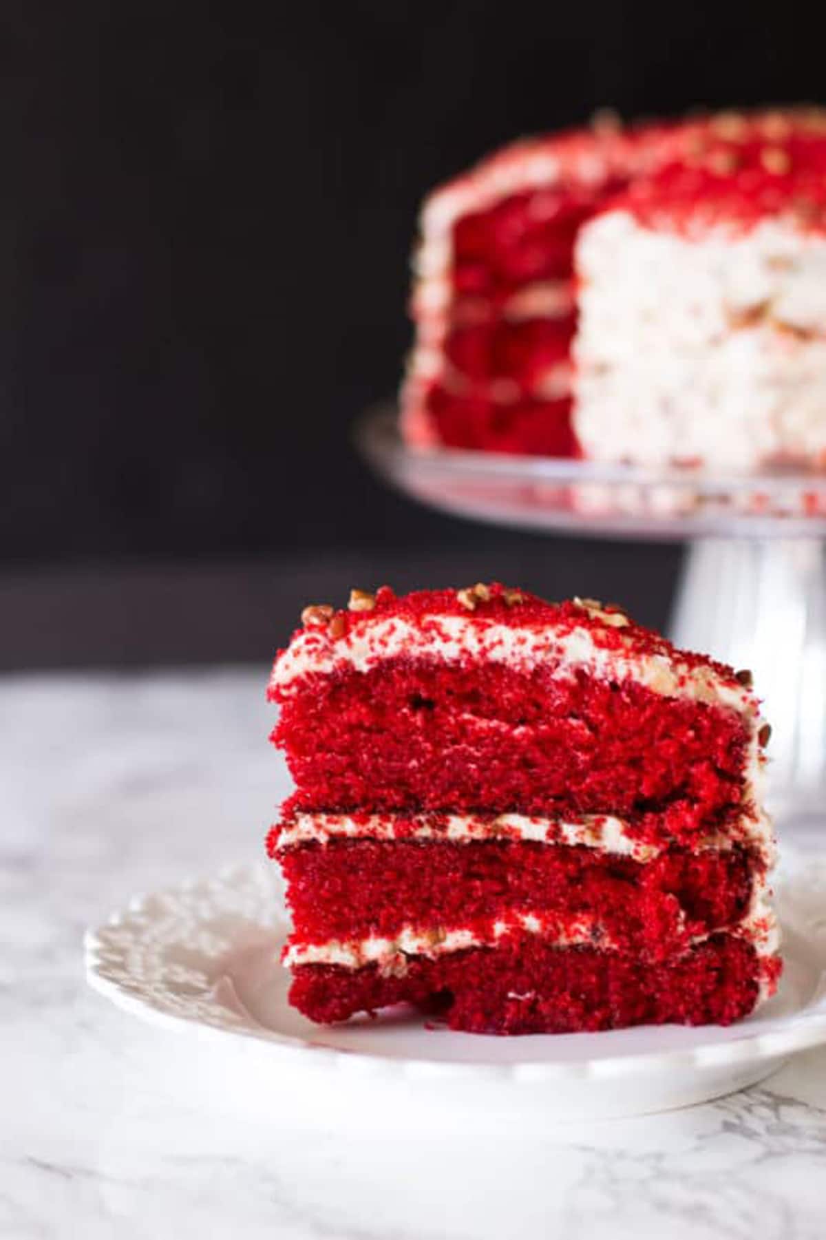 Piece of a 3 layered Red Velvet Cake sitting on a white lace plate, sliced Red Velvet Cake sitting on a glass cake dish.