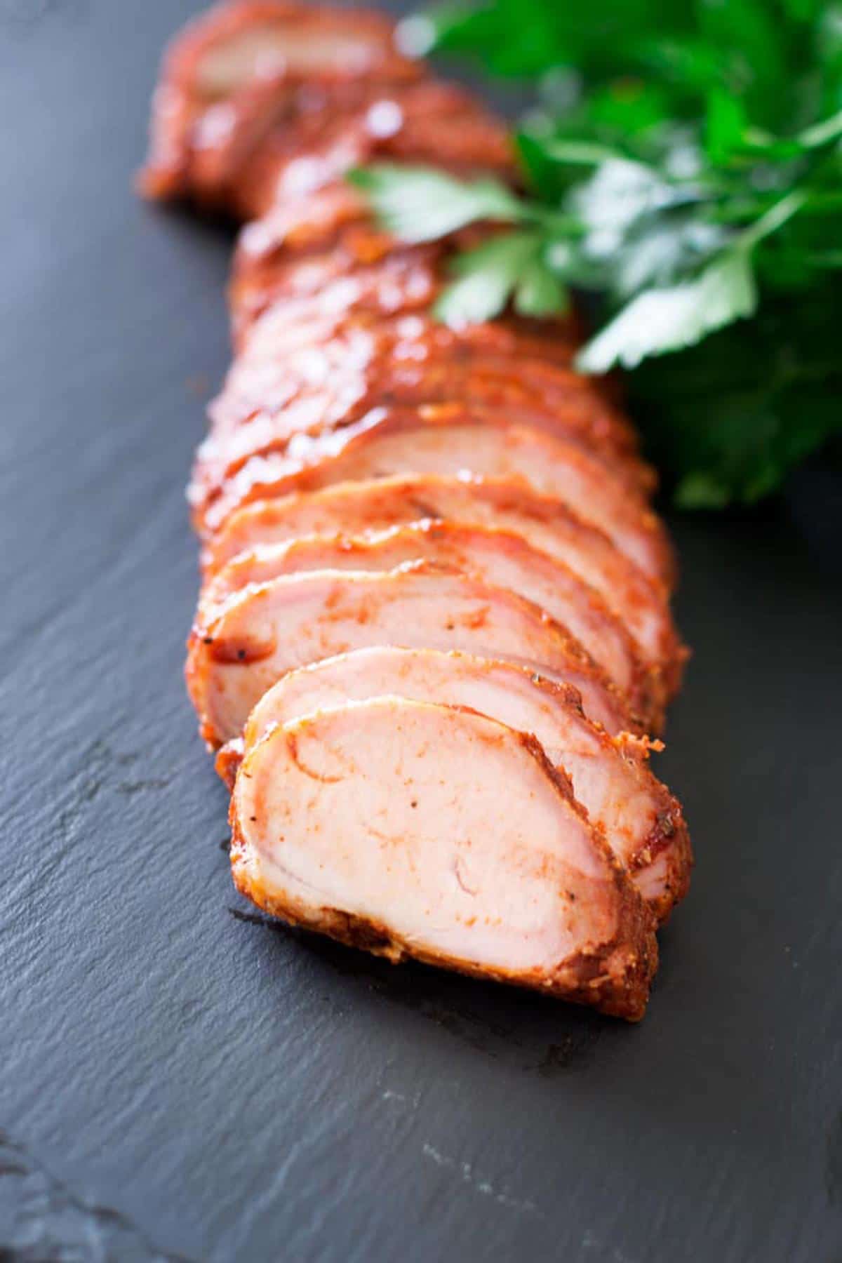 Sliced pork tenderloin covered with BBQ and on a black slate tray.