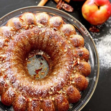 Apple pumpkin bundt cake sitting on a glass cake platter, 2 red apples with cinnamon stick and pecans on a black table.