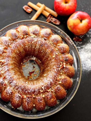 Apple pumpkin bundt cake sitting on a glass cake platter, 2 red apples with cinnamon stick and pecans on a black table.