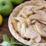 Pie shell containing apple pie filling, granny smith apples and peelings on table.