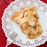 Slice of homemade apple pie in a white late plate sitting on a red table, fork on plate.