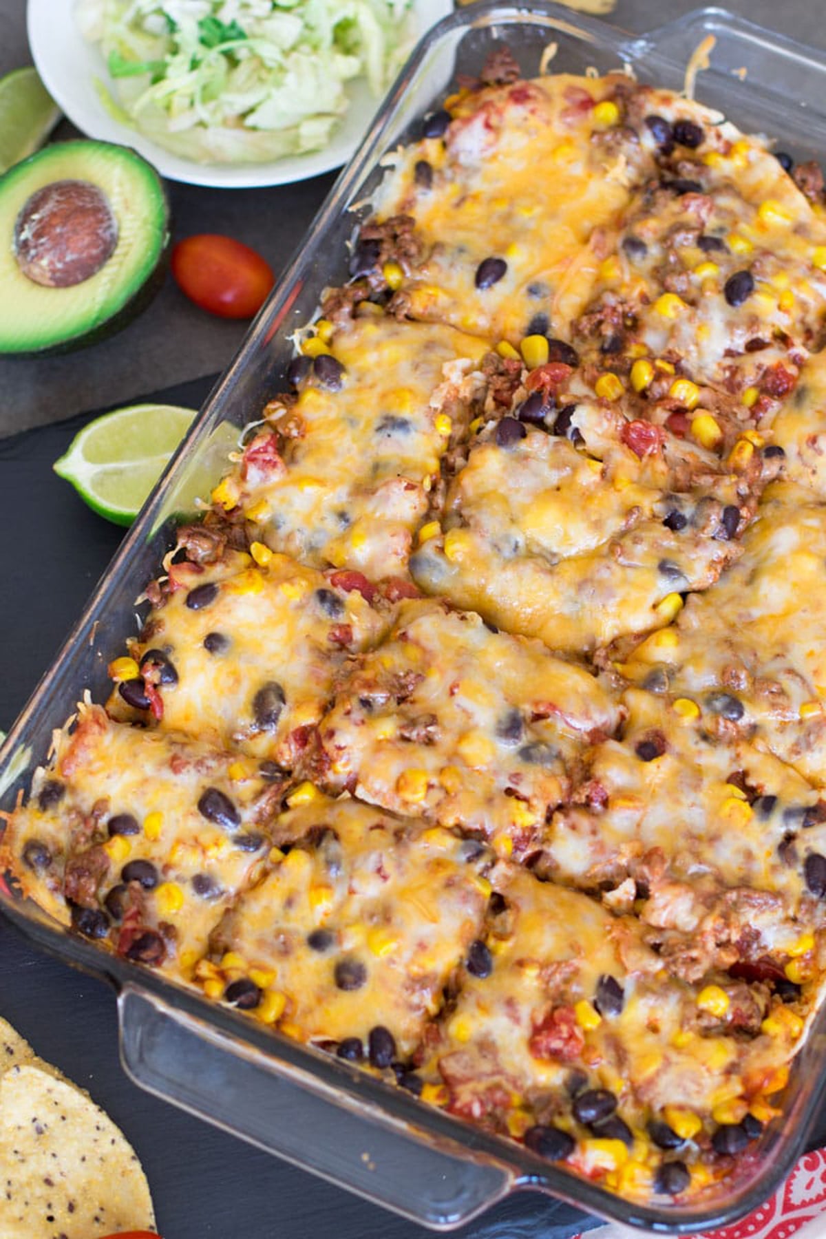 Beef enchilada casserole cut into 12 pieces, avocado, lime, and lettuce on table.