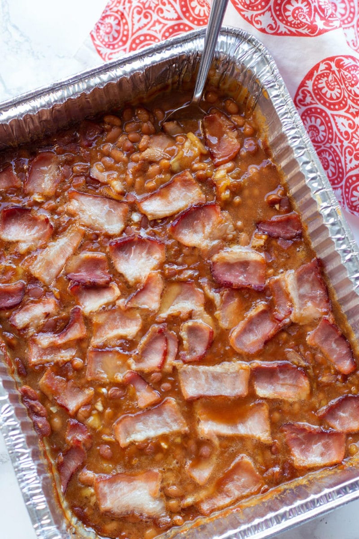 Pan containing baked beans cooked in a smoker topped with bacon. 
