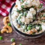 Spoonful of cauliflower and pea salad with cashews in a glass bowl sitting on a white gingham napkin, scattered peas and cashew nuts, silver spoon in bowl.