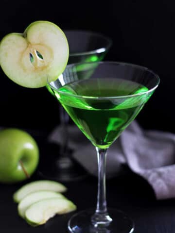 Two Martini glasses filled with a Green Apple Martini drink sitting on a black table, gray napkin and green apple slices in the background.