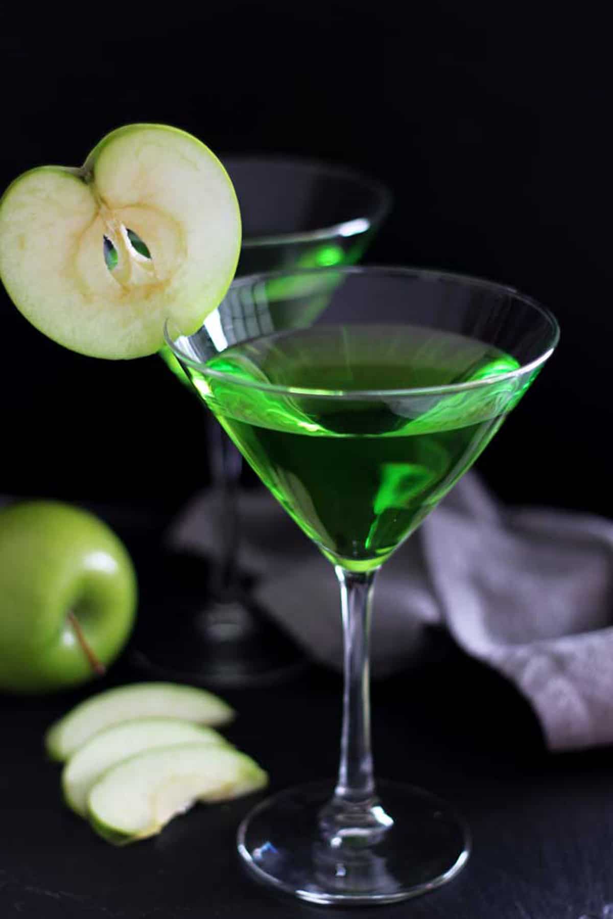 Two Martini glasses filled with a Green Apple Martini drink sitting on a black table, gray napkin and green apple slices in the background.