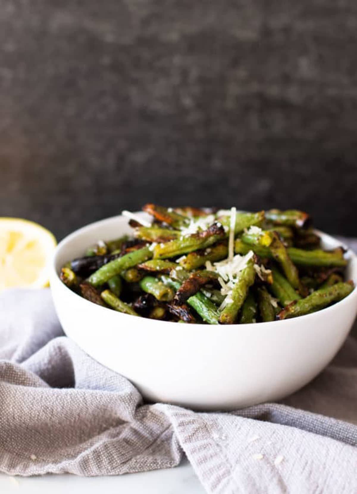 White bowl containing grilled green beans, lemon on table. 