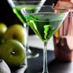 Two Martini glasses filled with a Green Apple Martini drink sitting on a black table, gray napkin and green apple in the background with a cocktail shaker.