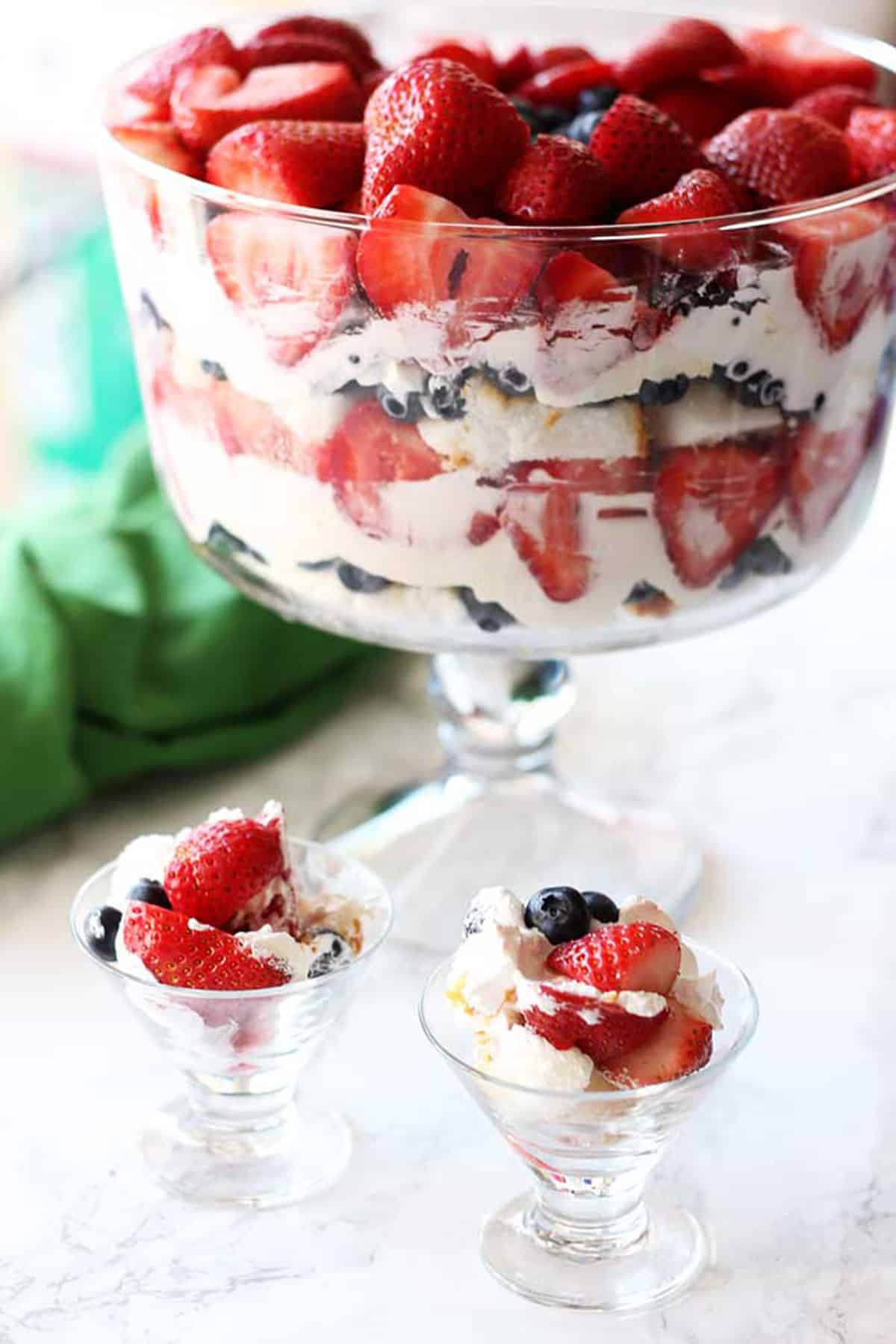 Trifle bowl containing cheesecake filling, angel food cake with strawberries and blueberries, 2 parfait cups will with trifle on counter.