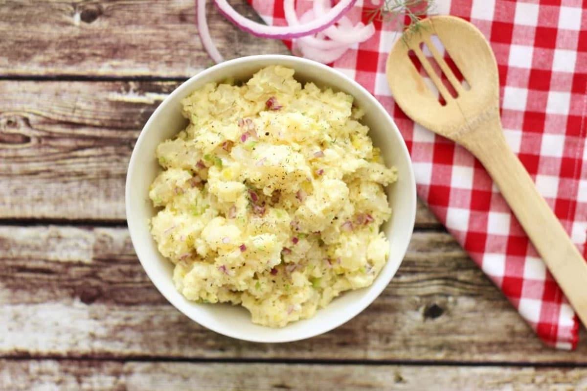 White bowl of potato salad sitting on a picnic table with a red and white check napkin, red onion ring and serving spoon.