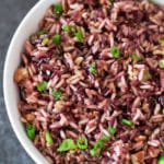 White bowl containing wild rice topped with scallions, pecans and dried cranberries on table.