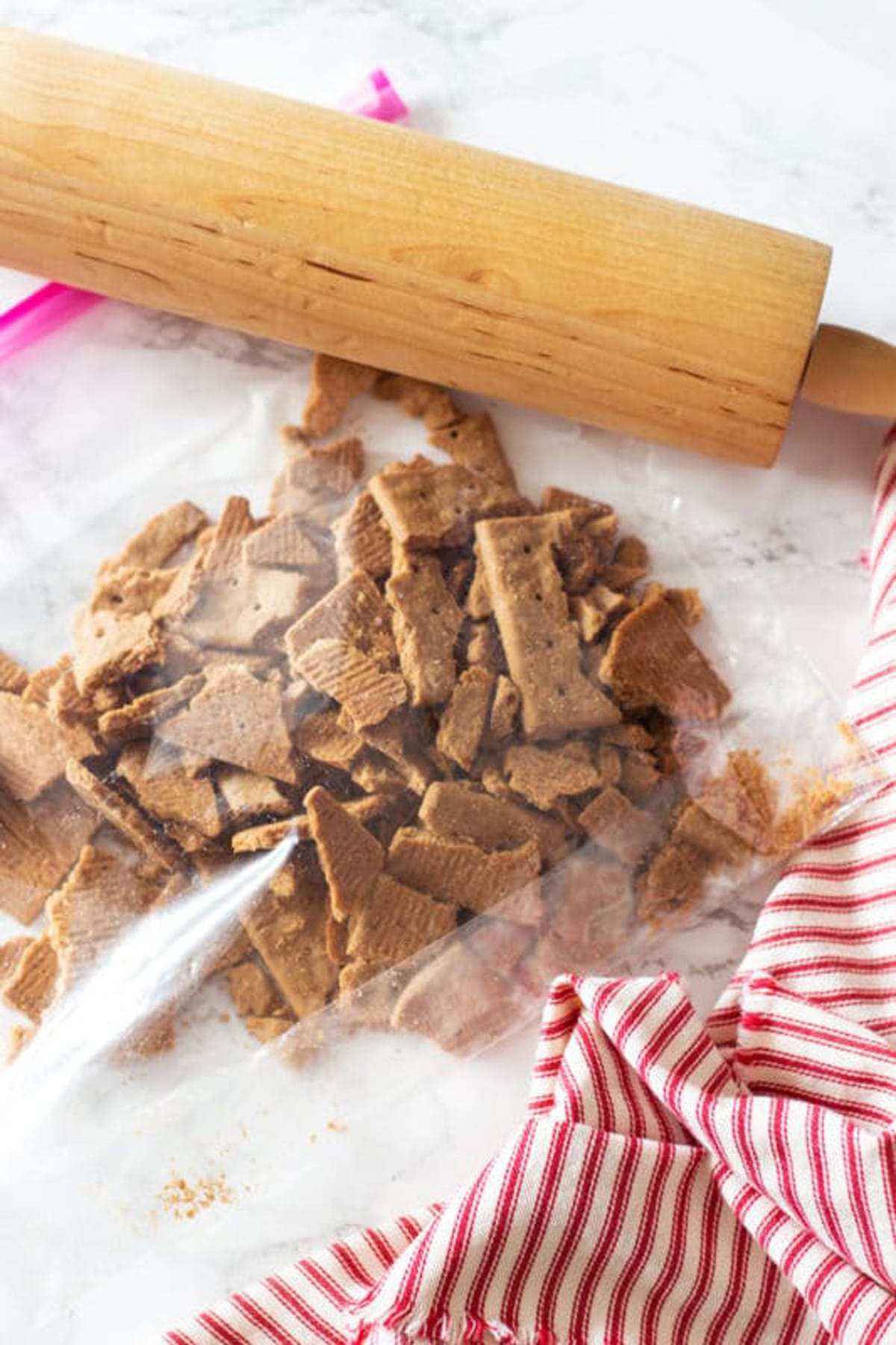 Ziplock bag containing crumbled graham crackers, rolling pin on table.