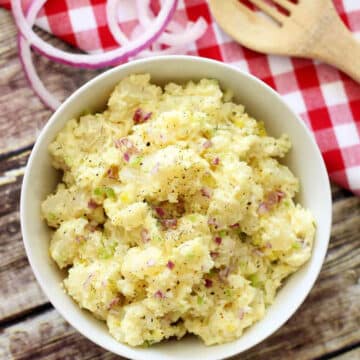 White bowl of Potato Salad sitting on a wooden table, gingham napkin with onion slices and a wooden spoon.