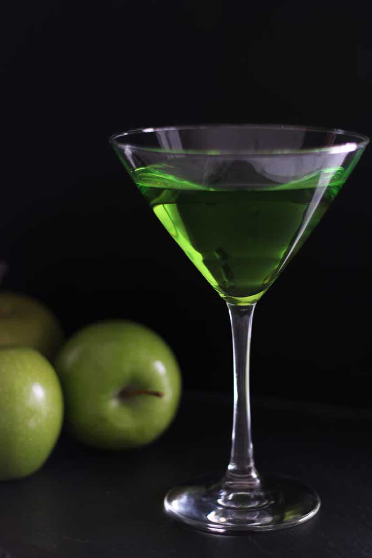 Martini glass filled with a Green Apple Martini Cocktail sitting on a black table, three green apples sitting on black table.