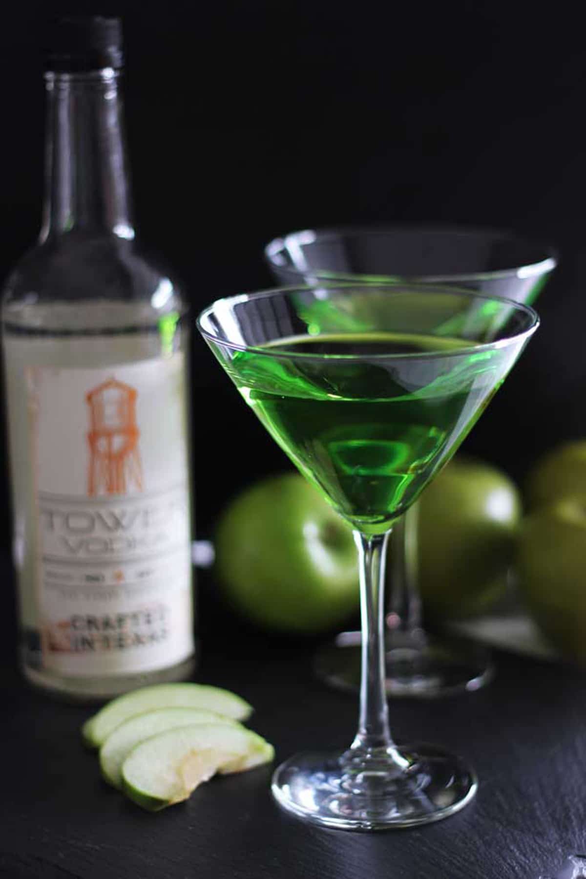 Two martini glasses filled with a Green Apple Martini cocktail sitting on a black table, Tower Vodka Bottle and green apples on the table.