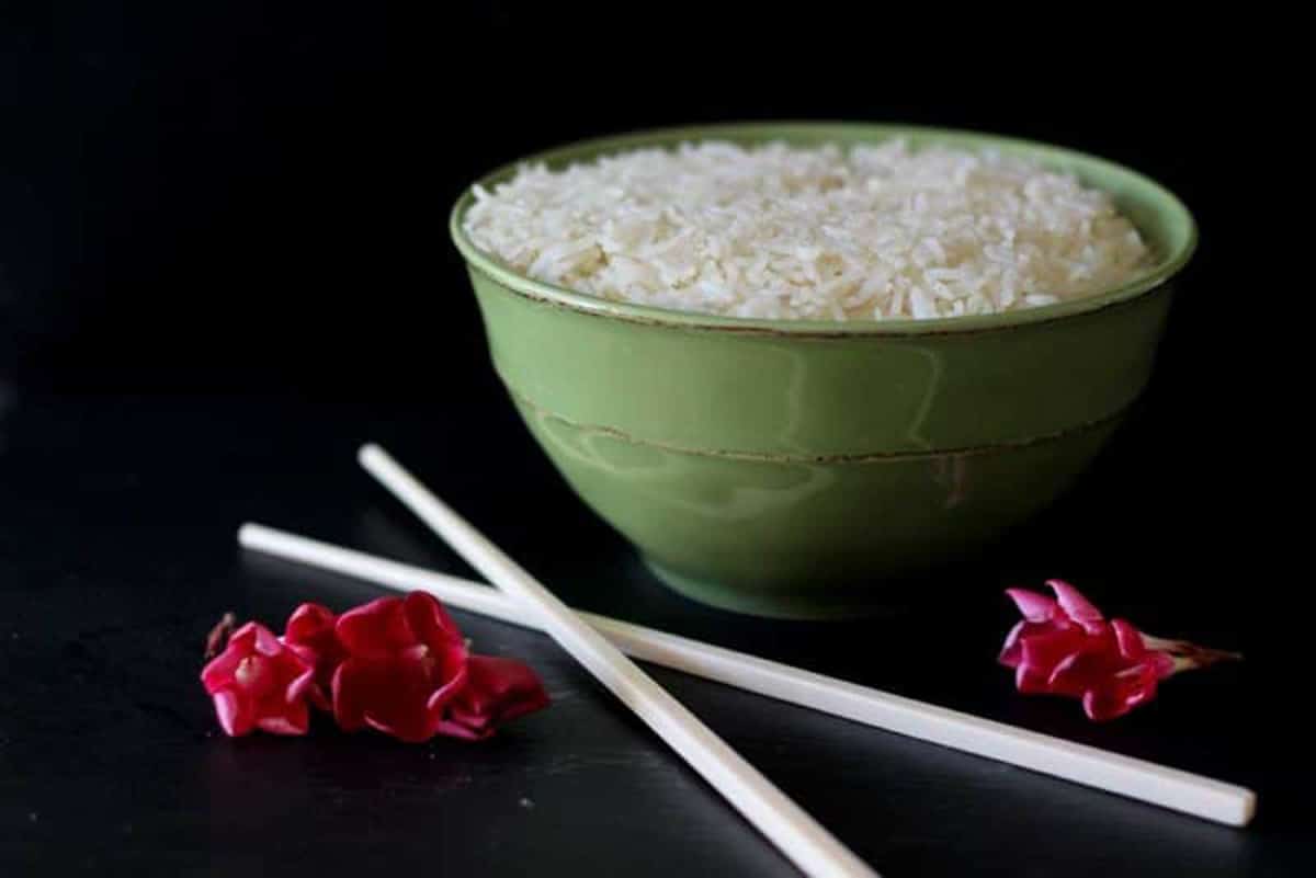 Green bowl sitting on a black table containing white rice, crossed chopsticks and 3 red flowers on table.