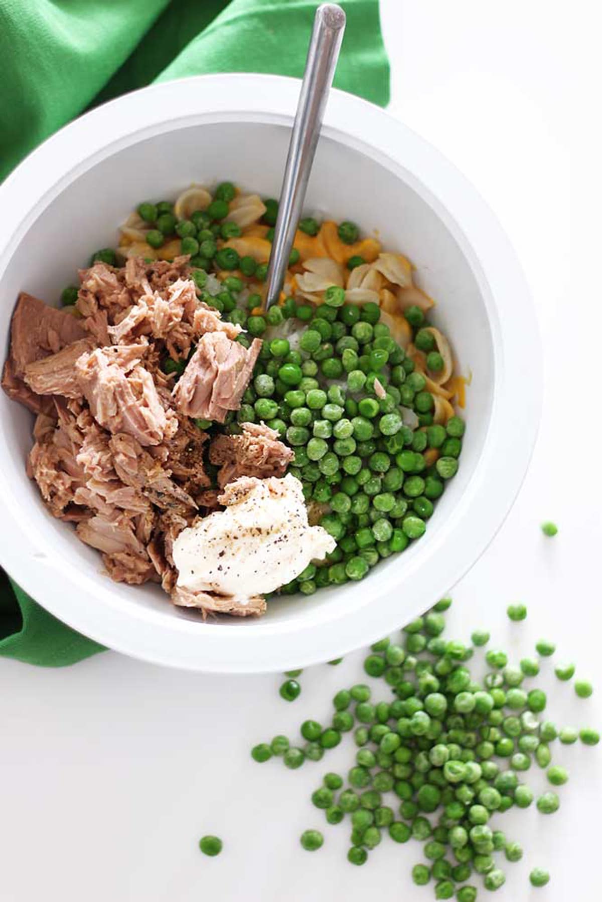 White mixing bowl with canned tuna, peas, shells and cheese and a silver spoon sitting on a white background with loose peas and a green napkin.